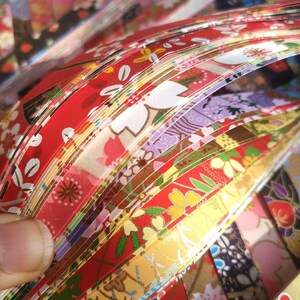 May include: A close-up of a stack of colorful origami paper strips. The strips feature various floral patterns in red, yellow, purple, and gold. The paper is fanned out, showcasing the different designs and colors.
