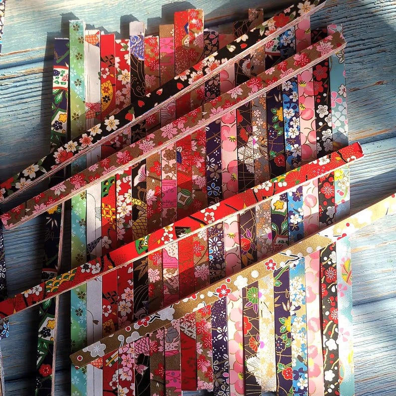 May include: A collection of colorful origami paper strips with floral and geometric patterns. The strips are arranged in a layered, angled display, showcasing a variety of colors including red, pink, black, and gold. The background is a light blue wooden surface.