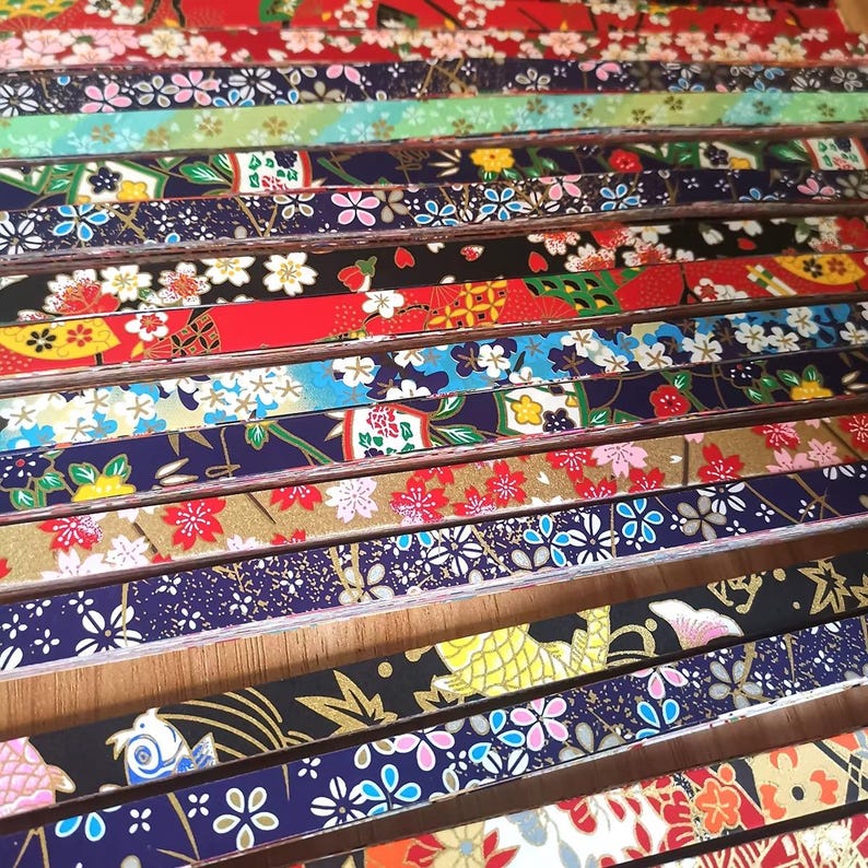May include: A collection of colorful Japanese origami paper strips. The strips feature various floral and geometric patterns in red, blue, gold, and white. The paper is arranged in parallel rows, showcasing the diverse designs.