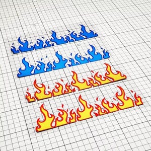 Puede incluir: Tres juegos de pegatinas con forma de llama en azul, amarillo y rojo. Cada juego tiene una combinación de colores diferente, con las llamas delineadas en un color contrastante. Las pegatinas están dispuestas en diagonal sobre un fondo de cuadrícula.