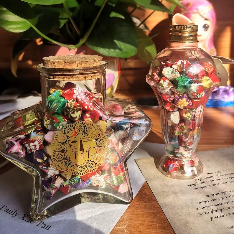 May include: Two glass containers filled with colorful origami stars. One is star-shaped with a cork lid, the other is a vase shape with a gold-colored cap. The containers sit on a table with a plant in the background.