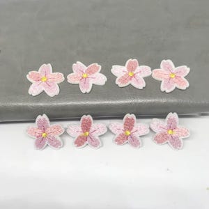 Può includere: Otto toppe floreali ricamate nei toni del rosa, bianco e giallo. I petali dei fiori sono dettagliati con cuciture e le toppe sono disposte su una superficie grigia e una superficie bianca.