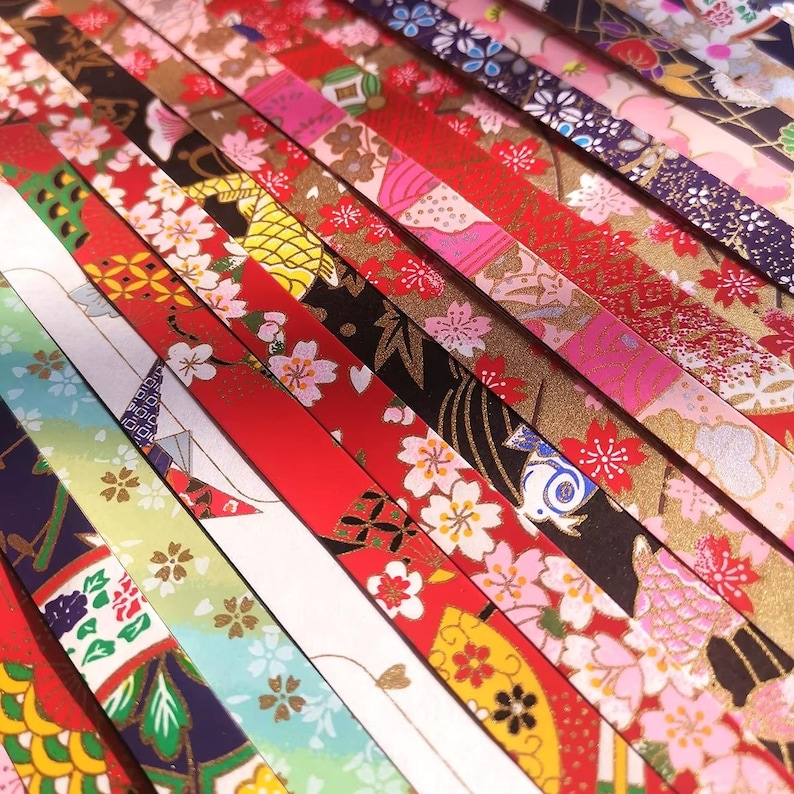 May include: A collection of colorful origami paper strips with various Japanese-inspired patterns. The strips feature floral designs, geometric shapes, and traditional motifs in red, gold, white, and black. The paper is arranged diagonally, showcasing the variety of patterns.