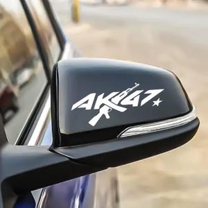 May include: A black car side mirror with a white graphic of an AK-47 assault rifle and the text "AK47". The design includes a star and is likely a decal or sticker for vehicle customization.