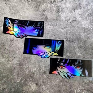 May include: Three holographic stickers featuring anime-style eyes with black hair and hands. The eyes have different colors, including purple, blue, and red. The stickers have a black background and a rainbow effect.