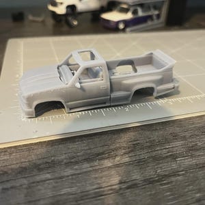 May include: A light grey model pickup truck with an open cab and bed. The truck lacks wheels and is positioned on a grey cutting mat with ruler markings. Other model vehicles are visible in the background.