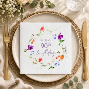 May include: A square white paper napkin featuring a colorful watercolor floral wreath surrounding the purple text "Marilyn's 90th birthday". The design includes various small flowers in shades of red, orange, blue, and purple with green leaves.