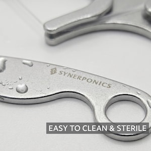 May include: A silver-coloured metal tool with a circular handle and a curved body, featuring the engraved text SYNERPONICS and a logo. The surface has small water droplets, and a dark rectangular label displays the text EASY TO CLEAN & STERILE.