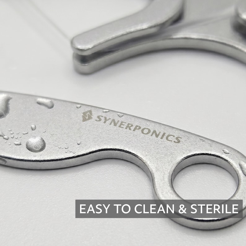 May include: A silver-coloured metal tool with a circular handle and a curved body, featuring the engraved text SYNERPONICS and a logo. The surface has small water droplets, and a dark rectangular label displays the text EASY TO CLEAN & STERILE.