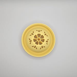 Op de afbeelding: Een geel, cirkelvormig decoratief object met een bloemmotief. Het midden van het object heeft een bloemvorm, omgeven door bladvormige uitsparingen. Het object is waarschijnlijk gemaakt van keramiek of een vergelijkbaar materiaal.
