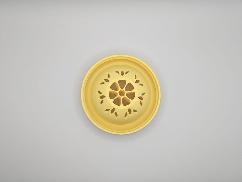 Op de afbeelding: Een geel, cirkelvormig decoratief object met een bloemmotief. Het midden van het object heeft een bloemvorm, omgeven door bladvormige uitsparingen. Het object is waarschijnlijk gemaakt van keramiek of een vergelijkbaar materiaal.