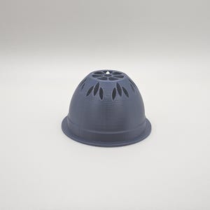 Op de afbeelding: Een donkergrijs, klokvormig object met een uitlopende basis en decoratieve uitsparingen. De bovenkant van het object heeft een reeks bloemblad-vormige openingen. Het object lijkt te zijn gemaakt van een vast materiaal, mogelijk plastic of keramiek.