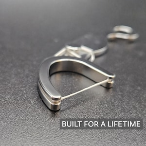 May include: A U-shaped silver-toned metal dental floss holder with a thin white string stretched across the opening. The device features a smooth, polished finish and is accompanied by a small metal ring attachment. The text BUILT FOR A LIFETIME is visible.