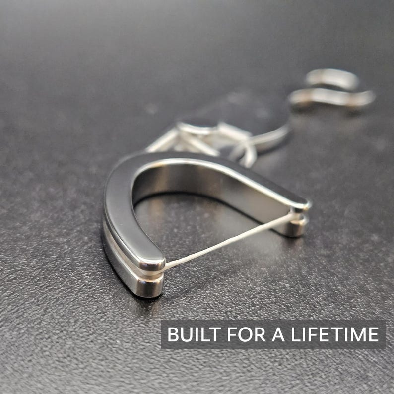 May include: A U-shaped silver-toned metal dental floss holder with a thin white string stretched across the opening. The device features a smooth, polished finish and is accompanied by a small metal ring attachment. The text BUILT FOR A LIFETIME is visible.