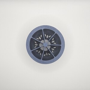 Op de afbeelding: Een cirkelvormig, blauw en grijs decoratief object met een citrusvruchtenontwerp. Het object is verdeeld in segmenten, elk met een reeks kleine, bladvormige uitsparingen. Het ontwerp is gecentreerd op een witte achtergrond.