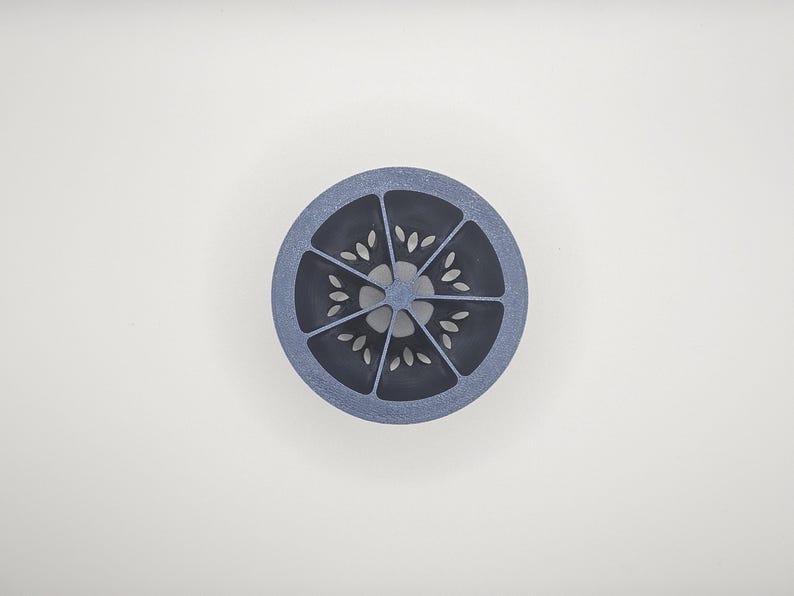 Op de afbeelding: Een cirkelvormig, blauw en grijs decoratief object met een citrusvruchtenontwerp. Het object is verdeeld in segmenten, elk met een reeks kleine, bladvormige uitsparingen. Het ontwerp is gecentreerd op een witte achtergrond.