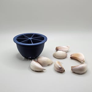 Op de afbeelding: Een donkerblauwe knoflookpers met een gesegmenteerde bovenkant en een afgeronde basis. Verschillende knoflookteentjes liggen verspreid rond de pers op een lichtgrijze ondergrond. De pers heeft kleine gaatjes aan de onderkant.