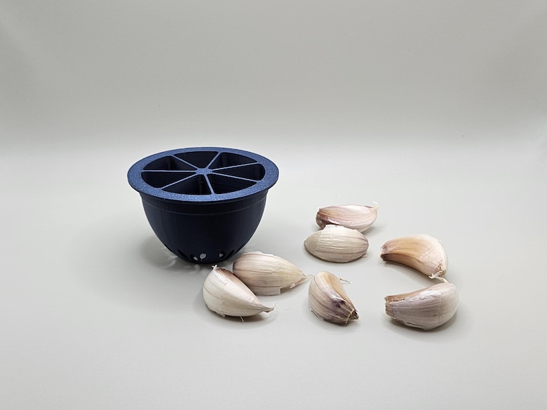 Op de afbeelding: Een donkerblauwe knoflookpers met een gesegmenteerde bovenkant en een afgeronde basis. Verschillende knoflookteentjes liggen verspreid rond de pers op een lichtgrijze ondergrond. De pers heeft kleine gaatjes aan de onderkant.