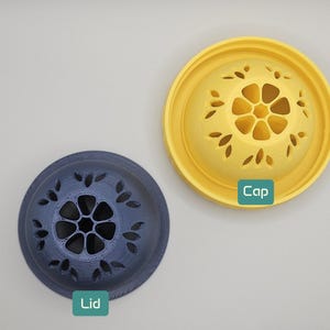 Op de afbeelding: Bovenaanzicht van twee decoratieve deksels. E&eacute;n is donkerblauw met een bloemmotief en het woord "Lid" in een groene doos. De andere is geel met een bloemmotief en het woord "Cap" in een groene doos.