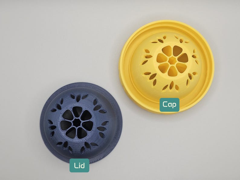 Op de afbeelding: Bovenaanzicht van twee decoratieve deksels. E&eacute;n is donkerblauw met een bloemmotief en het woord "Lid" in een groene doos. De andere is geel met een bloemmotief en het woord "Cap" in een groene doos.