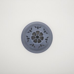 Op de afbeelding: Een cirkelvormig, grijs decoratief object met een bloemenontwerp. Het object heeft bloembladvormige uitsparingen langs de rand en een centraal bloemachtig patroon. Het is gecentreerd op een witte achtergrond.