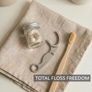 May include: A small clear glass jar containing organic silk floss with a silver metal lid, a silver metal floss holder with a circular handle, and a light brown wooden toothbrush. The text on the jar reads Organic Silk Floss.