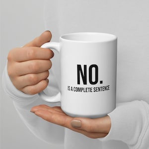 May include: White ceramic mug with the text "NO. IS A COMPLETE SENTENCE" in black. The mug has a curved handle and a cylindrical shape. The mug is held in a person's hands.
