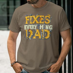 Könnte beinhalten: Braunes T-Shirt mit dem Text "FIXES EVERYTHING DAD" in Gelb und Weiß. Der Text ist mit Werkzeugen in den Buchstaben gestaltet. Das Shirt hat einen Rundhalsausschnitt.