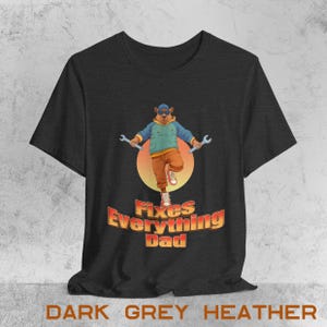 Könnte beinhalten: Dunkelgraues meliertes T-Shirt mit einem Cartoon-Bären, der eine Sonnenbrille trägt und Schraubenschlüssel hält. Der Bär steht vor einem Sonnenuntergang, darunter der Text "Fixes Everything Dad".