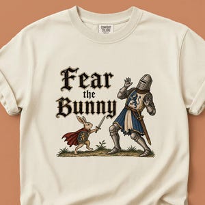 May include: A cream-coloured t-shirt featuring a graphic illustration of a small rabbit wearing a red cape and holding a sword, facing a knight in full armour. The text "Fear the BUNNY" is printed above the figures in a bold, black gothic font.