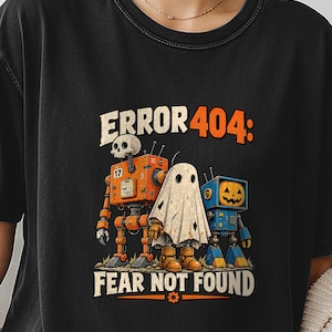 Op de afbeelding: Een zwart t-shirt met een grafische print met de tekst ERROR 404: FEAR NOT FOUND. Het ontwerp toont een oranje robot met een schedelhoofd, een wit spookfiguur en een blauwe robot met een pompoenhoofd die naast elkaar staan.