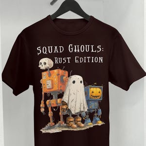 May include: Dark brown t-shirt featuring the text "SQUAD GHOULS: RUST EDITION." The graphic depicts three robot-like figures: a skull, a ghost, and a jack-o'-lantern, all with a Halloween theme.
