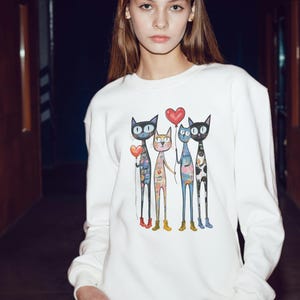 Może przedstawiać: Biała bluza z kolorową ilustracją czterech stylizowanych kotów. Każdy kot ma unikalny wzór i trzyma balon w kształcie serca. Koty stoją razem, tworząc fantazyjny i artystyczny design.