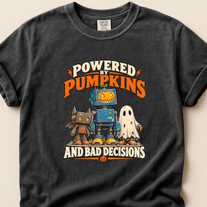 May include: A charcoal grey t-shirt featuring a graphic of a bat-like robot, a blue robot with a pumpkin head, and a ghost. The text reads "POWERED BY PUMPKINS AND BAD DECISIONS" in orange and white lettering, with a small pumpkin icon at the bottom.