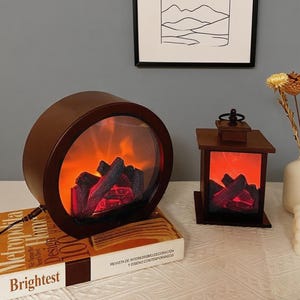 Puede incluir: Dos faroles decorativos eléctricos con forma de chimenea. Uno es un farol redondo, marrón oscuro, con una parte delantera transparente, que muestra una llama naranja brillante y leños falsos. El otro es un farol rectangular más pequeño.