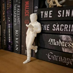 May include: A white figurine of a person reading, leaning against a stack of books. The figure is wearing a helmet and trousers, and is holding a book. The books have titles like "Falling Shadows" and "The Sini".