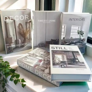 以下が含まれることがあります： 「DECOR」、「TIMELESS LIVING」、「INTERIORS」、「STILL」などのタイトルが書かれた白い表紙のインテリア関連書籍のコレクション。インテリアデザインの写真が掲載されており、白い面に配置されています。