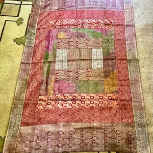 May include: A red, gold, and green patterned throw blanket with a fringed edge. The blanket is made of a soft, woven fabric and has a patchwork design.