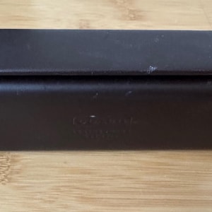 May include: A rectangular, dark brown leather case with a hinged lid. The edges are stitched with white thread. The case is resting on a light brown wooden surface. The case appears to be for eyeglasses.