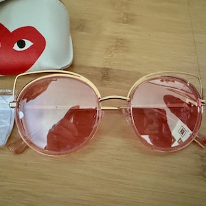 Puede incluir: Gafas de sol redondas con montura dorada y lentes rosas. Las gafas presentan un diseño único con una fina montura dorada que se extiende por encima de las lentes. Se ve una funda blanca con un diseño de corazón y ojo rojo.