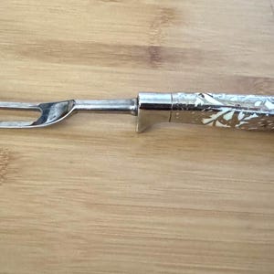 Op de afbeelding: Een zilveren trancheervork met een decoratief handvat. De handgreep heeft een gegraveerd bloemmotief met witte accenten. De vork met twee tanden is gemaakt van gepolijst metaal. De trancheervork ligt op een houten oppervlak.