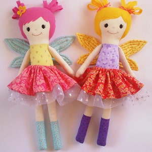 May include: Two handmade fabric dolls dressed as fairies. The doll on the left has pink hair, a yellow bodice, and a pink and red skirt with a tulle overlay. The doll on the right has yellow hair, a purple bodice, and a red and orange skirt with a tulle overlay.