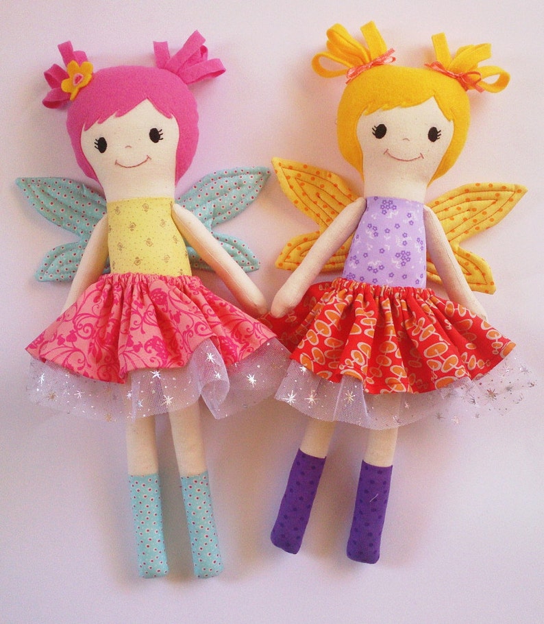 May include: Two handmade fabric dolls dressed as fairies. The doll on the left has pink hair, a yellow bodice, and a pink and red skirt with a tulle overlay. The doll on the right has yellow hair, a purple bodice, and a red and orange skirt with a tulle overlay.