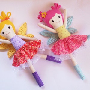 May include: Two handmade fabric fairy dolls with wings. One doll has yellow hair, a purple and orange dress, and yellow wings. The other doll has pink hair, a yellow and pink dress, and blue wings.