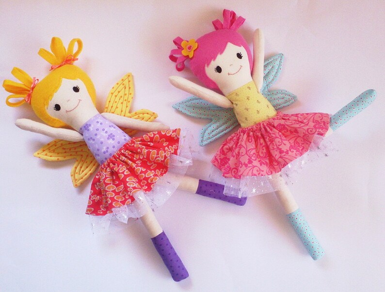 May include: Two handmade fabric fairy dolls with wings. One doll has yellow hair, a purple and orange dress, and yellow wings. The other doll has pink hair, a yellow and pink dress, and blue wings.
