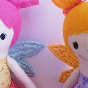 May include: Two handmade fabric dolls with wings. The doll on the left has pink hair and blue and white wings. The doll on the right has yellow hair and orange and yellow wings.