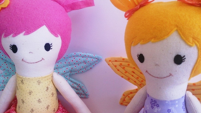 May include: Two handmade fabric dolls with wings. The doll on the left has pink hair and blue and white wings. The doll on the right has yellow hair and orange and yellow wings.