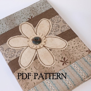 以下が含まれることがあります： 茶色とベージュの布製のジャーナルカバーで、大きな白い花のアップリケと中央に茶色のボタンが付いています。カバーはさまざまな模様の布で作られており、下部に「PDF PATTERN」という文字が印刷されています。