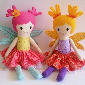 May include: Two handmade fabric fairy dolls with wings. One doll has pink hair, a yellow bodice, and a pink skirt. The other doll has yellow hair, a purple bodice, and a red skirt.