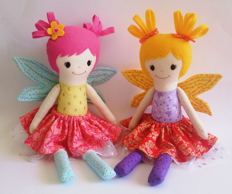 May include: Two handmade fabric fairy dolls with wings. One doll has pink hair, a yellow bodice, and a pink skirt. The other doll has yellow hair, a purple bodice, and a red skirt.